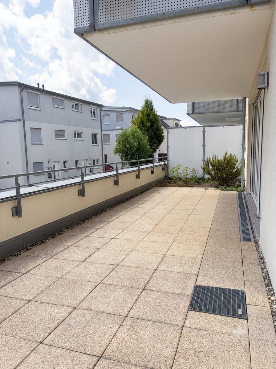 Blick über die Balkonterrasse in die Gegenrichtung, mit überdachtem Bereich, bepflanztem Beet an der Hauswand und Ausblick auf die gegenüberliegenden Wohnhäuser