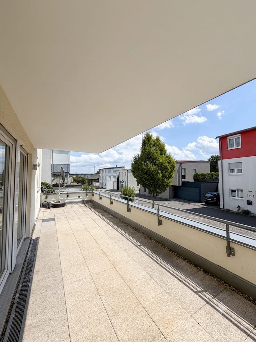 Überdachte Balkonterrasse mit hellen Bodenplatten und Metallgeländer, links eine bodentiefe Schiebetür zur Wohnung, dahinter ein Baum und die Nachbarbebauung