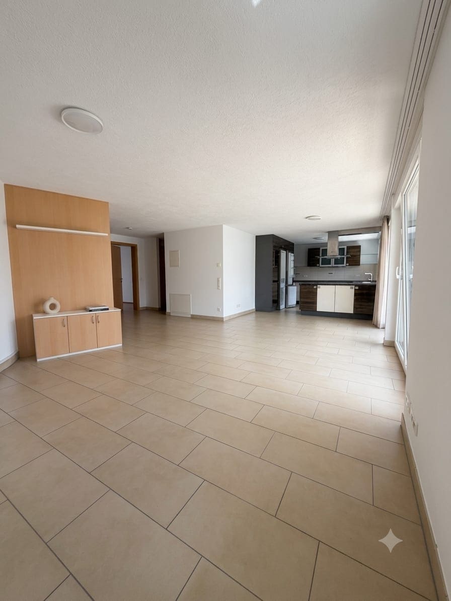 Leerer, heller Wohn- und Essbereich mit Fliesenboden, Einbauschrank an der linken Wand und offener Küche am hinteren Ende, rechts bodentiefe Fenster zur Terrasse